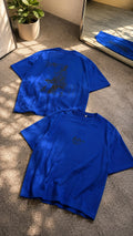 Two bright blue short-sleeve T-shirts laid flat on a carpeted floor, with one shirt showing a dark graphic print on the back and the other showing a small dark chest logo. A potted plant and mirror are visible in the room setting.