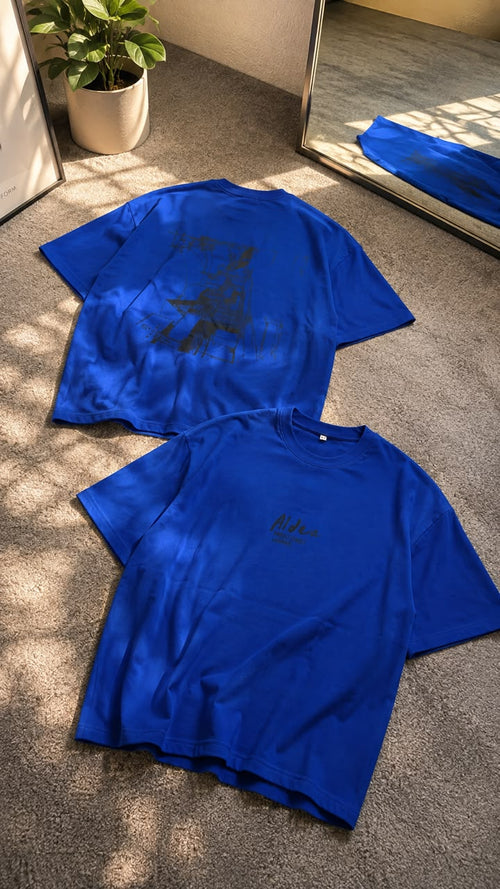 Two bright blue short-sleeve T-shirts laid flat on a carpeted floor, with one shirt showing a dark graphic print on the back and the other showing a small dark chest logo. A potted plant and mirror are visible in the room setting.